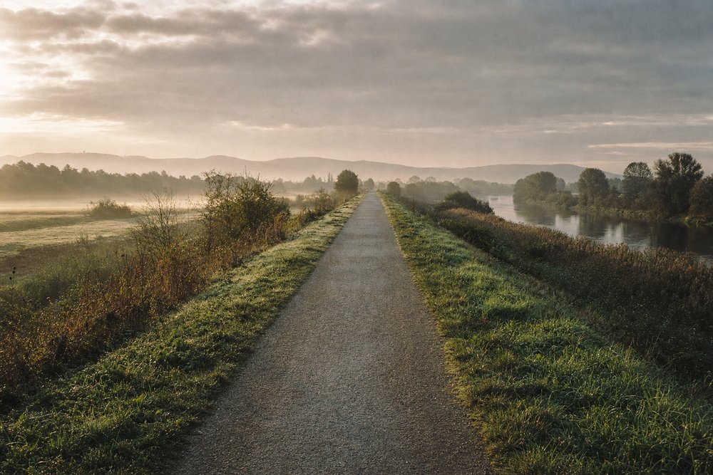 Flacher, autofreier Weg auf einer ehemaligen Bahntrasse durch das Tal der Wetterau im Morgennebel, mit einem Fluss zur Seite und den Hügeln des Taunus im Hintergrund